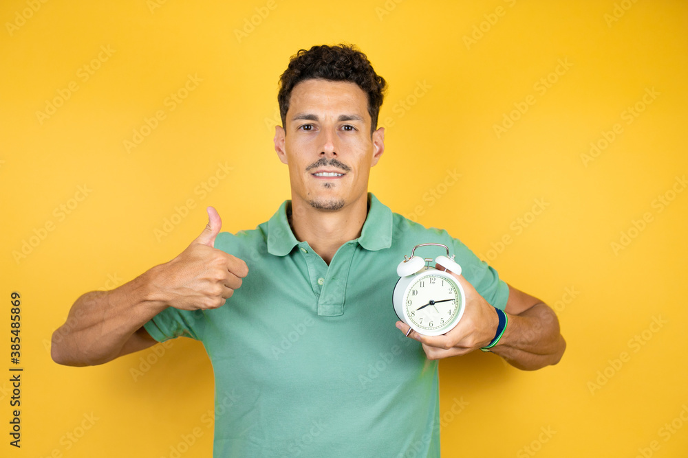 Young handsome man wearing green casual t-shirt over isolated yellow background pointing and holding a clock Young handsome man wearing green casual t-shirt over isolated yellow background pointing and holding a clock