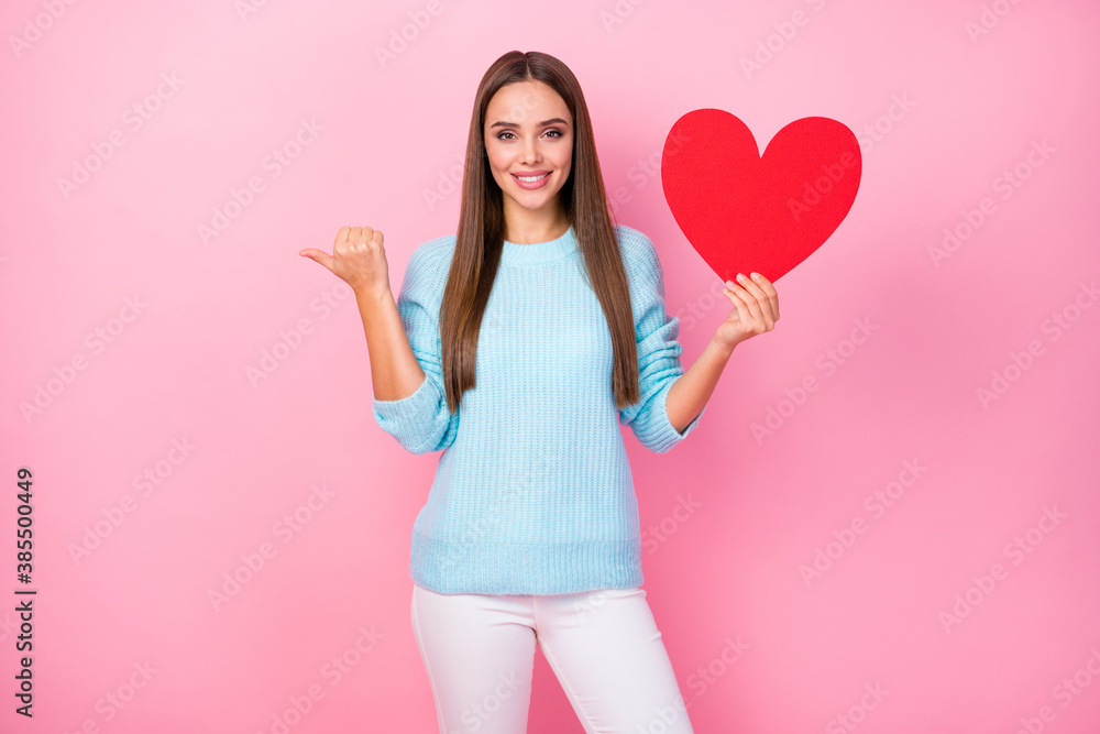 Photo of attractive lady hold big paper red heart direct thumb finger empty space advice creative postcard idea wear knitted blue sweater white pants isolated pastel pink color background Photo of attractive lady hold big paper red heart direct thumb finger empty space advice creative postcard idea wear knitted blue sweater white pants isolated pastel pink color background
