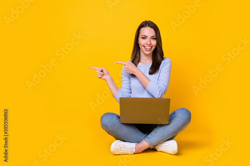 Full length photo of positive girl sit floors legs crossed work laptop point index finger advertise promo wear purple violet sweater denim jeans isolated bright shine color background photo