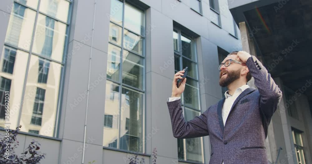 Front view of likable in high spirits successful adult bearded office worker in glasses which is surprised from good news on phone screen while standing near modern building Front view of likable in high spirits successful adult bearded office worker in glasses which is surprised from good news on phone screen while standing near modern building
