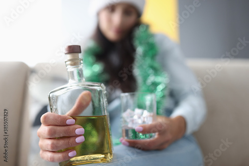 Glass bottle with alcohol close up. Woman in tinsel sit on couch and hold bottle of whiskey and glass of ice.