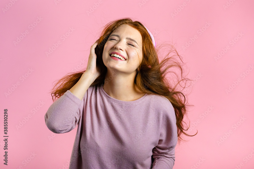Fototapeta premium Attractive woman with red hair with headphones on pink studio background. Cute girl portrait. Music, radio, happiness, freedom, youth concept.