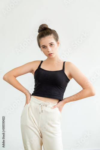 Home outfit. Casual sportive look. Pretty young woman in black sleeveless top white trousers and bun hairstyle holding hands on hip looking at camera isolated on neutral. Fitness clothes