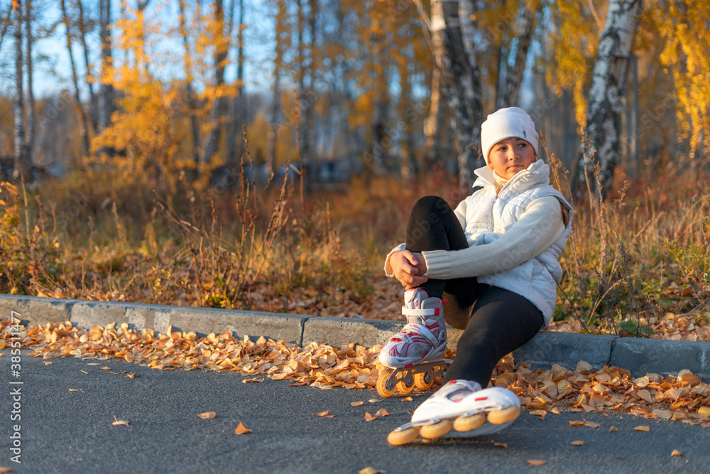 A girl in roller skates is sitting on concrete curb at the park and look away. A young figure skater is resting. Autumn evening. The girl is wearing a white hat, sweater, jacket and black leggings A girl in roller skates is sitting on concrete curb at the park and look away. A young figure skater is resting. Autumn evening. The girl is wearing a white hat, sweater, jacket and black leggings