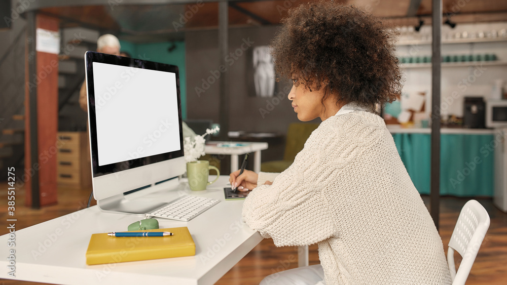 A good looking dark-skinned and fashionably dressed young woman sitting at a table in front of a PC drawing with a graphics tablet