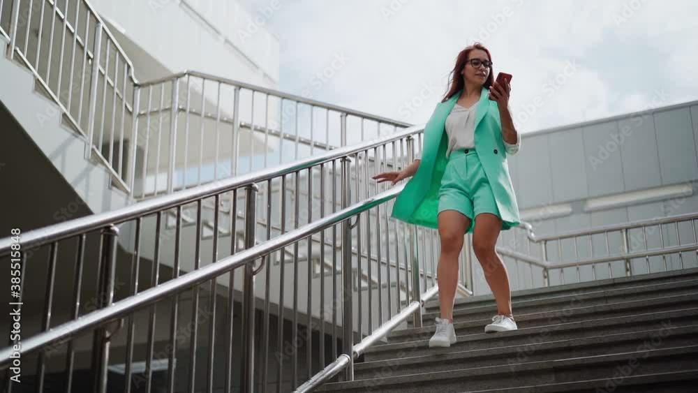 A beautiful happy woman in glasses and fashionable clothes turquoise color with a mobile phone in hand is going down the stairs business center, smiling and laughing with joy of success. Slow motion A beautiful happy woman in glasses and fashionable clothes turquoise color with a mobile phone in hand is going down the stairs business center, smiling and laughing with joy of success. Slow motion