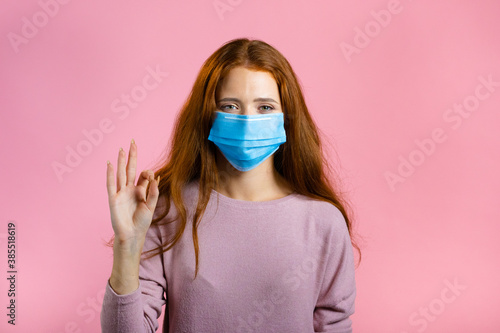 Happy woman in medical mask on pink background. Lady with long red hair shows Ok gesture. Winner. Success. Body language.