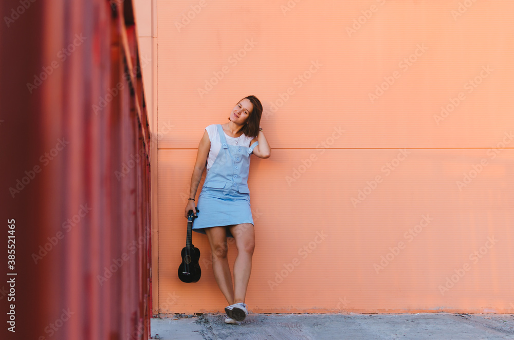 Fototapeta premium Young beautiful woman playing ukulele and having fun at the background of a coral wall