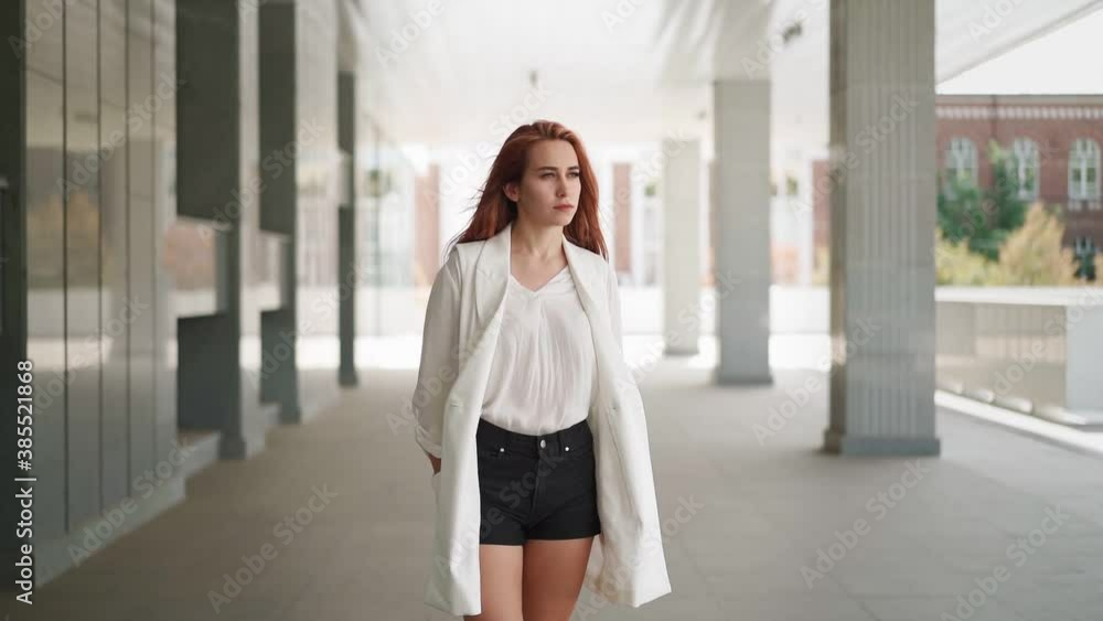Beautiful modern business woman with long red hair in trendy white coat stands near an office building. Confident female entrepreneur. Attractive successful lady in fashionable clothes. Slow motion Beautiful modern business woman with long red hair in trendy white coat stands near an office building. Confident female entrepreneur. Attractive successful lady in fashionable clothes. Slow motion