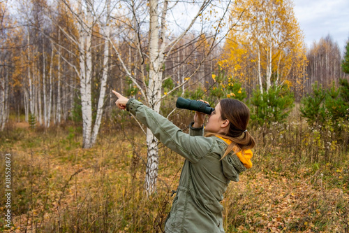 Young woman with binoculars watching birds and pointing in the direction of the bird in the autumn forest. Scientific research