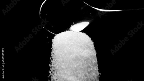White cane sugar falling down from teaspoon isolated on black background slow motion. Excess sugar leads to obesity, cardio-vascular diseases. Eat less sugar to reduce diabetes risk, healthy lifestyle