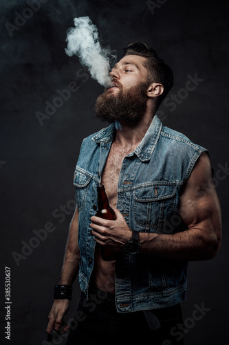 Cool and haired guy with naked torso and huge biceps posing holding bottle in arm and smoking vape in dark background.