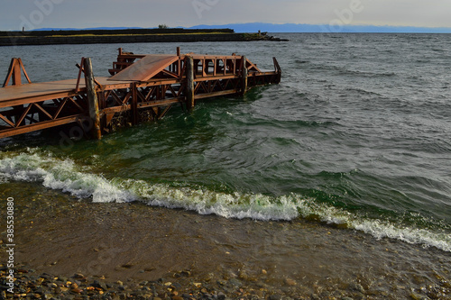 Wallpaper Mural Old rusty iron pier, red orange bright dock construction on sandy stone shore of Lake Baikal in green water with waves. Blue mountains on horizon. Bolshoe Goloustnoe Torontodigital.ca