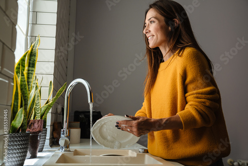 Beautiful middle-aged woman smiling while washing dishes at home