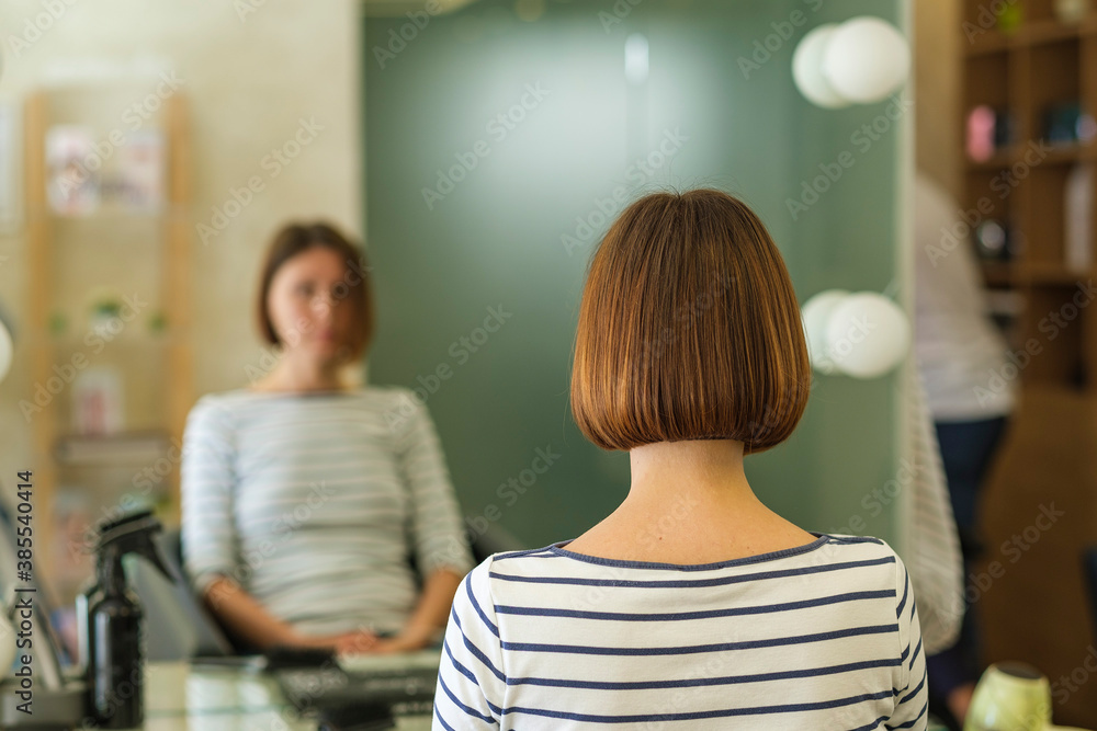 Young woman had her hair cut and styled in a beauty salon. She is sitting facing the mirror, and from behind we can see a straight line of her brown hair reaching up to the neck Young woman had her hair cut and styled in a beauty salon. She is sitting facing the mirror, and from behind we can see a straight line of her brown hair reaching up to the neck