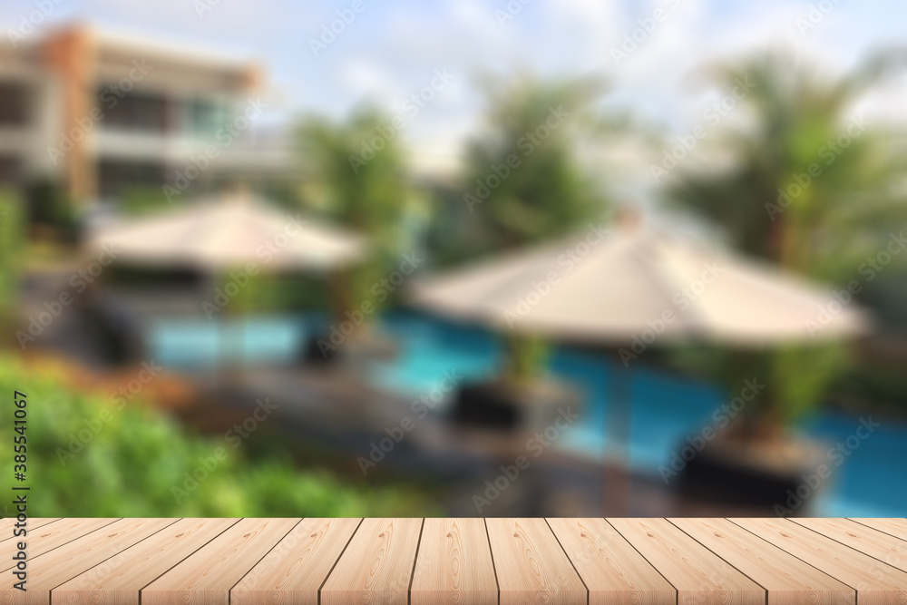 Empty wood plank table top with outdoor pool, trees, umbrellas and building with blur background - For product display.