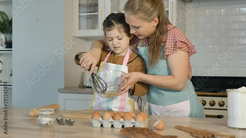 Careful mother teaches little daughter to whisk albuminous in glass bowl at table with ingredients for pie in light kitchen