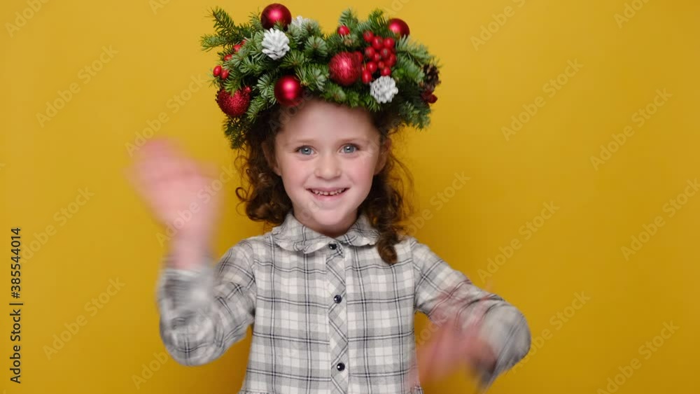 Portrait of cute little girl kid in traditional Christmas wreath, waves palm in hello gesture, greets friends, isolated on yellow studio background. Happy New Year celebration merry holiday concept Portrait of cute little girl kid in traditional Christmas wreath, waves palm in hello gesture, greets friends, isolated on yellow studio background. Happy New Year celebration merry holiday concept