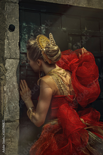 Beautiful young blonde woman wearing an elegant red and gold princess gown and golden jewels, looking through the key hole of a dungeon door