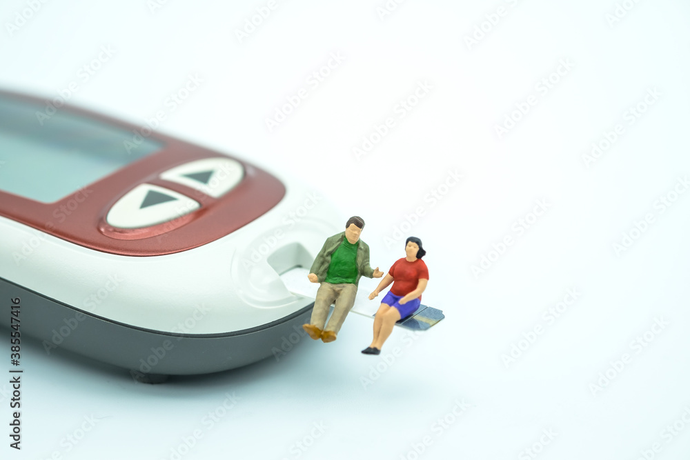 Fat man and woman miniature people figure sitting on test strip with Glucose meter for check blood sugar level on white background using as diabetes, glycemia, health care and people concept.