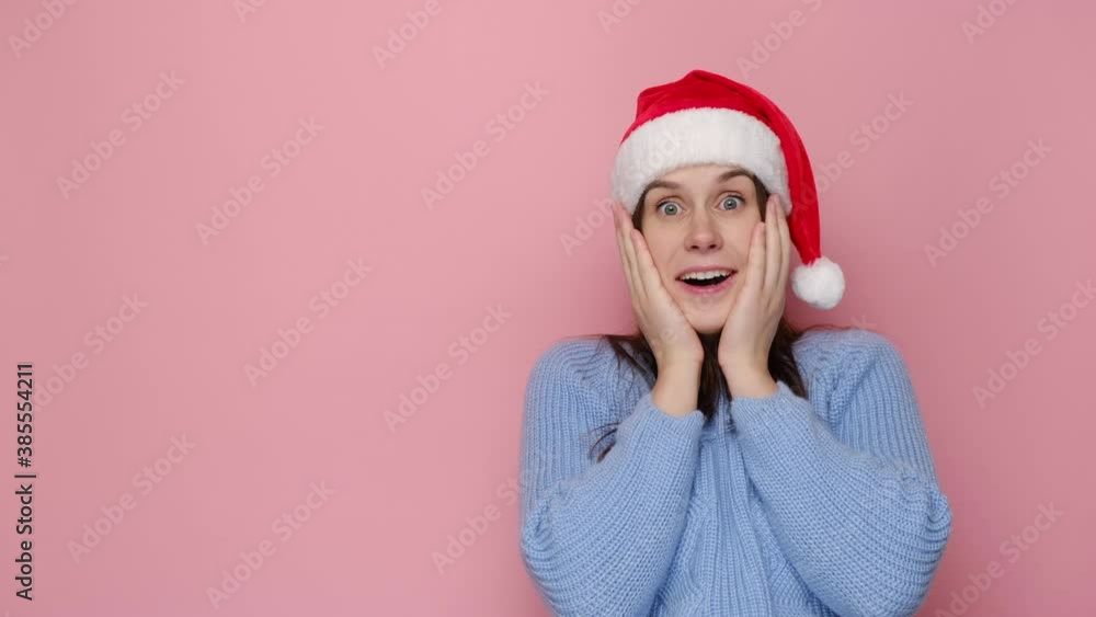 Emotional smiling young woman in Christmas red hat with shocked expression keeps palms on cheek, wears winter cozy sweater, waits for New Year, isolated over pink studio background. Xmas is coming Emotional smiling young woman in Christmas red hat with shocked expression keeps palms on cheek, wears winter cozy sweater, waits for New Year, isolated over pink studio background. Xmas is coming