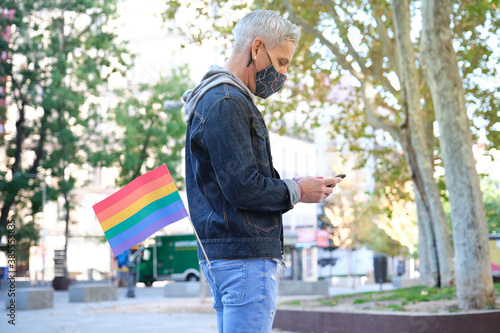 Young caucasian homosexual man wearing face mask with an LGBT flag in his pocket chatting on his smartphone. LGBT pride celebration in pandemic times. © Ladanifer