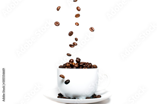 Roasted coffee beans falling into a coffee cup on a saucer with a white background, close-up, coffee wallpaper.