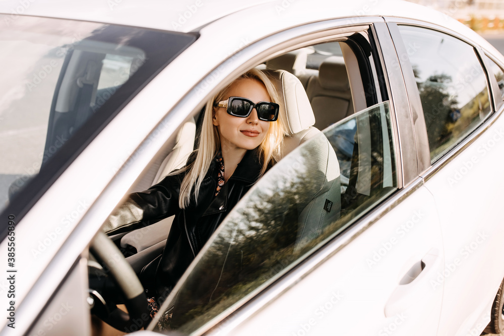 Young blonde woman driving a car, smiling, wearing black sunglasses and leather jacket.