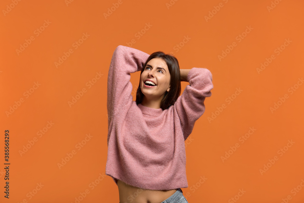 Im not afraid of problems. Portrait of laughing young female keeps hands behind head, enjoys leisure time. Positive emotions. Wears in pink knitted sweater isolated over orange wall