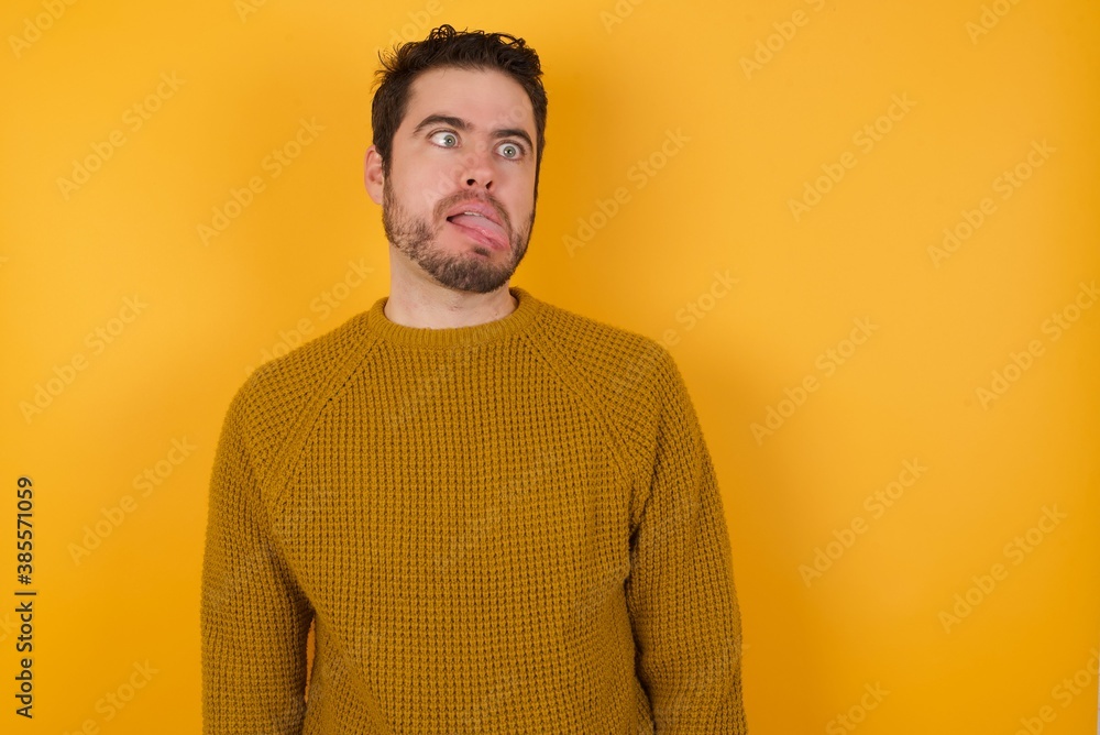 Young man wearing casual sweater and over isolated yellow background showing grimace face crossing her eyes and showing tongue. Being funny and crazy Young man wearing casual sweater and over isolated yellow background showing grimace face crossing her eyes and showing tongue. Being funny and crazy