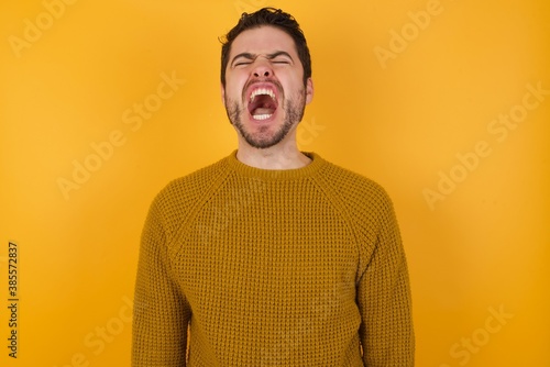 Young man wearing casual sweater and over isolated yellow background angry and mad screaming frustrated and furious, shouting with anger. Rage and aggressive concept.