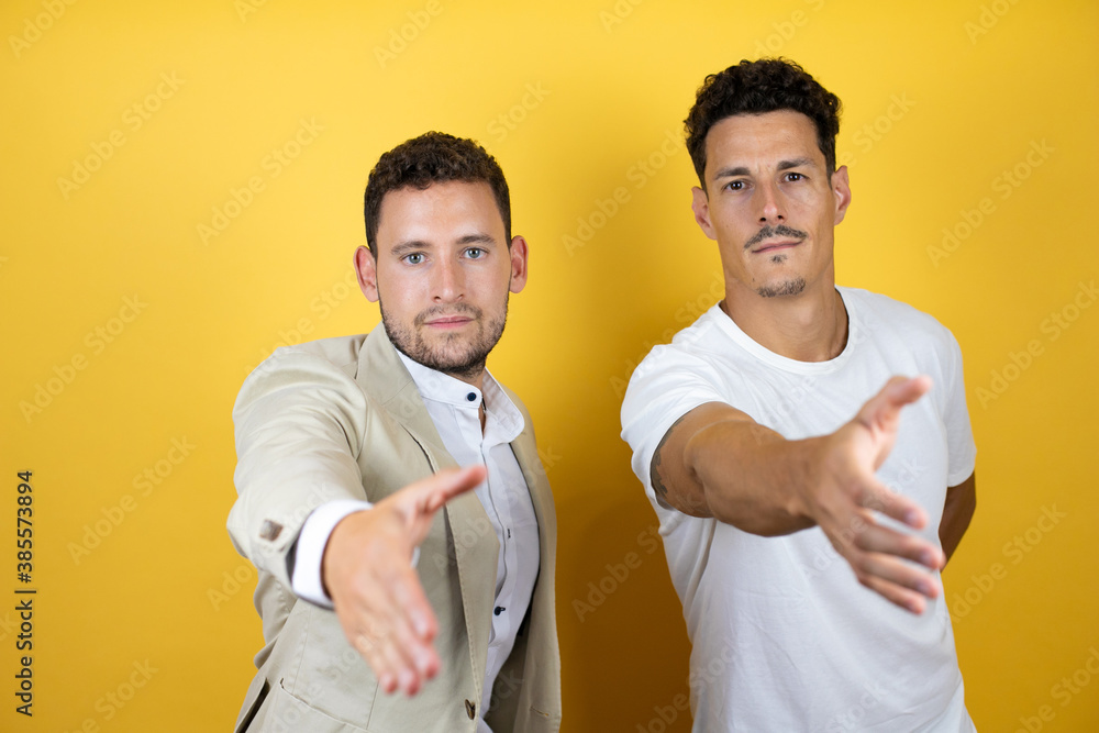 Young gay couple of two men wearing casual clothes over isolated yellow background smiling friendly offering handshake as greeting and welcoming Young gay couple of two men wearing casual clothes over isolated yellow background smiling friendly offering handshake as greeting and welcoming