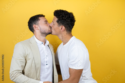 Young gay couple of two men wearing casual clothes over isolated yellow background Standing with smile on face kissing
