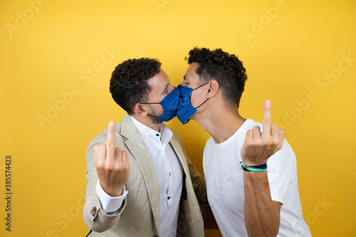 Young gay couple of two men wearing casual clothes and mask over isolated yellow background kissing and showing middle finger doing fuck you bad expression, provocation and rude attitude