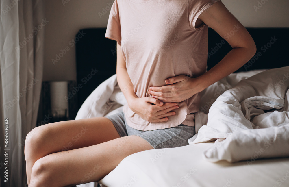 Young woman suffering from strong abdominal pain, casual style indoor shoot. Female sitting on bed in morning Young woman suffering from strong abdominal pain, casual style indoor shoot. Female sitting on bed in morning