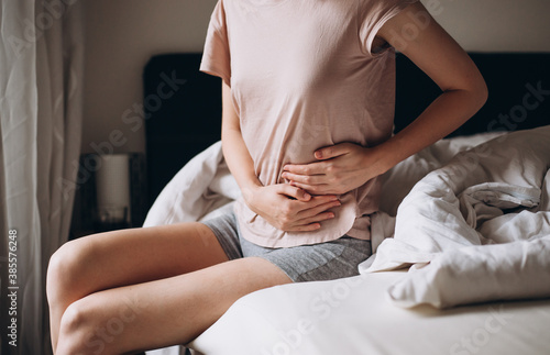 Young woman suffering from strong abdominal pain, casual style indoor shoot. Female sitting on bed in morning