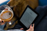 A woman reads from her e-book while enjoying a glass of beer on a couch, with her dog on it