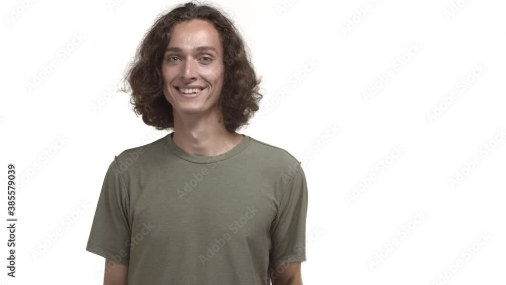 Attractive young hipster guy with wavy messy hairstyle, wearing t-shirt, waving at camera with friendly smile, saying hello, greeting someone, standing over white background Attractive young hipster guy with wavy messy hairstyle, wearing t-shirt, waving at camera with friendly smile, saying hello, greeting someone, standing over white background