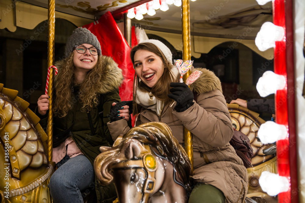 Two female friends riding on the carousel with horses at the winter festival