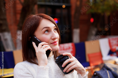 Smiling brunette woman talking on a smartphone while having her takeaway coffee. Receiving good news.