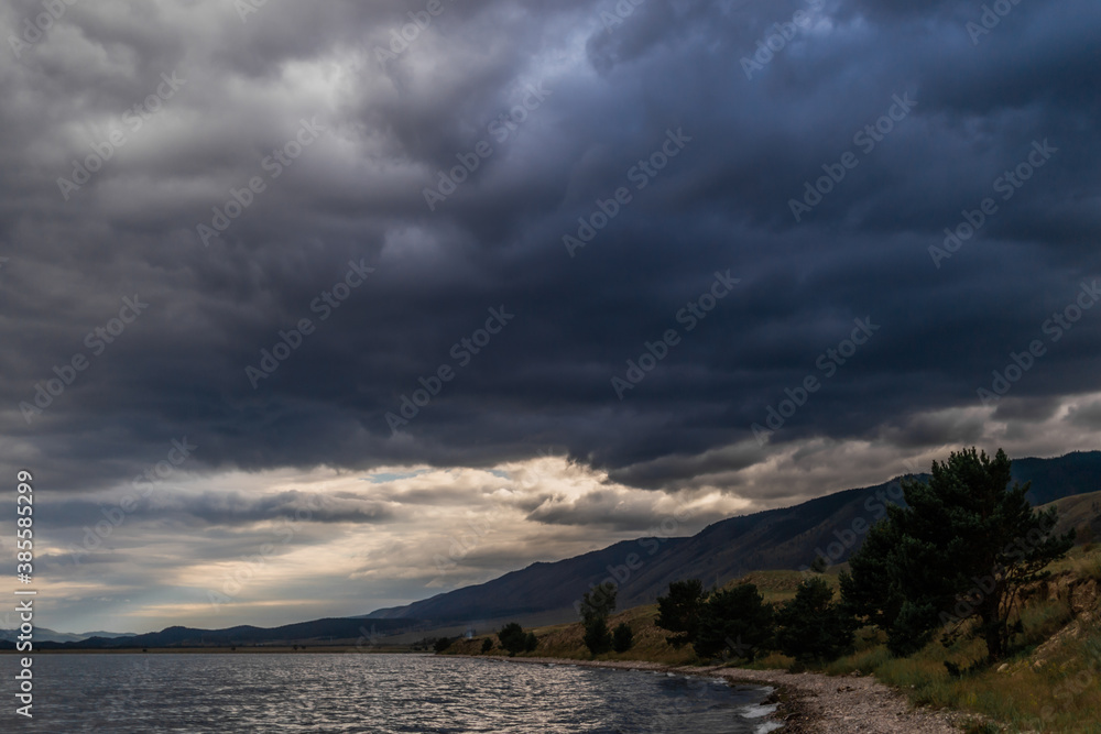 Pebbles shore with green trees of blue Lake Baikal, mountains on the horizon, dark clouds. Evening landscape