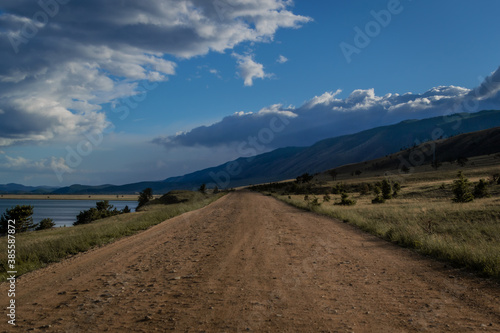 Road on green grassy with trees shore of lake Baikal with dark blue mountains. Summer landscape in sunset light
