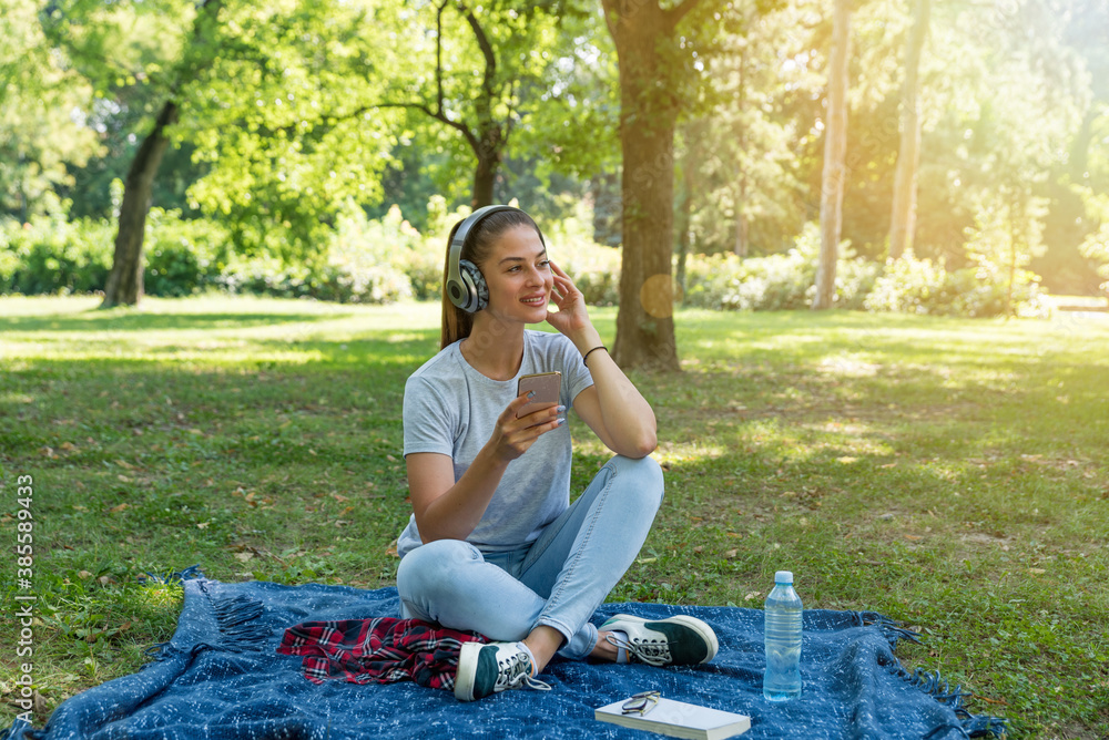 Young happy woman sitting in the park surfing the song play list in her phone and listening the music on the wireless headphones Young happy woman sitting in the park surfing the song play list in her phone and listening the music on the wireless headphones
