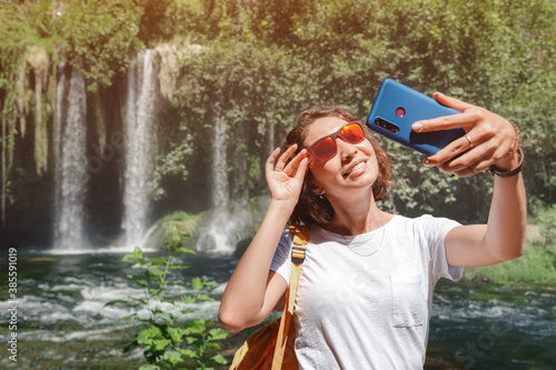 Fototapeta Naklejka Na Ścianę i Meble -  Asian female travel blogger takes a selfie photo against the backdrop of a powerful waterfall in the wild jungle. Social networks and the Internet during hiking concept