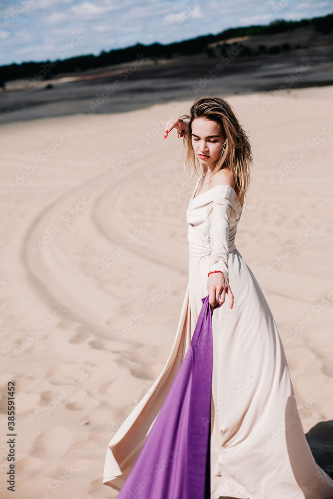 Fototapeta premium A young, slender girl in a beige dress with purple cloth in her hands posing in the desert in the wind
