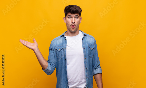 young handsome man looking surprised and shocked, with jaw dropped holding an object with an open hand on the side photo