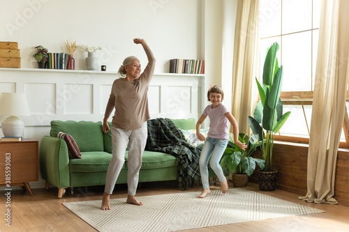 Overjoyed barefoot elderly female grandma or babysitter dancing with cute girl on floor carpet at cozy home. Excited grandmother having fun with small child granddaughter in living room. photo