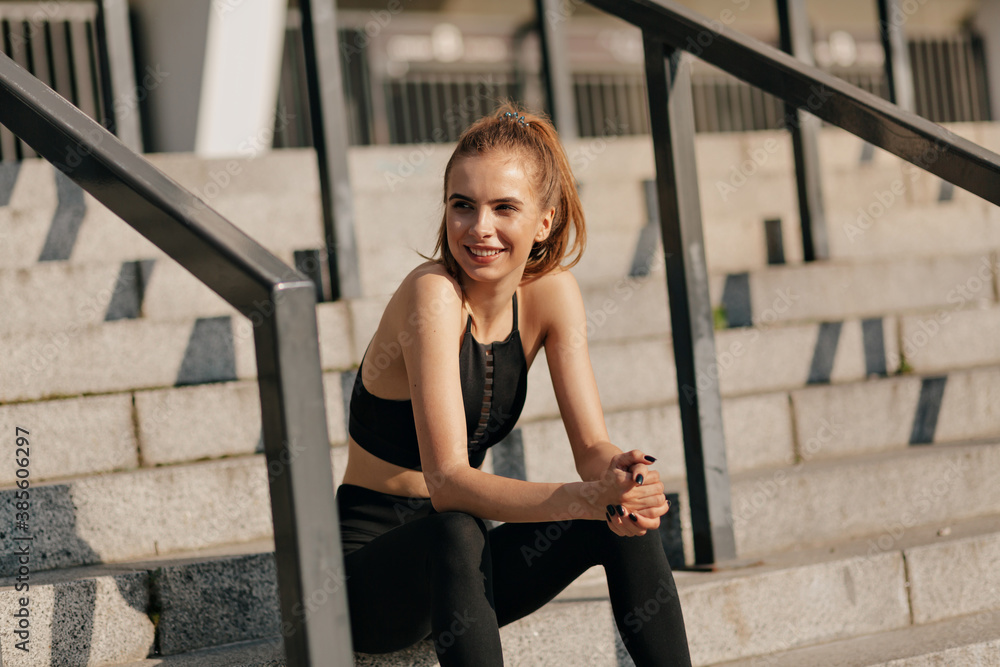 Happy smiling girl on outside stairs looking away with smile in sport uniform. Outdoor photo of beautiful white girl enjoying training at stadium.