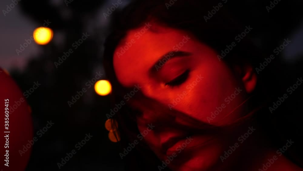 Beautiful young hispanic woman performer and entertainer looking serious and confident at night under dramatic red street lighting with colorful balloons. Beautiful young hispanic woman performer and entertainer looking serious and confident at night under dramatic red street lighting with colorful balloons.