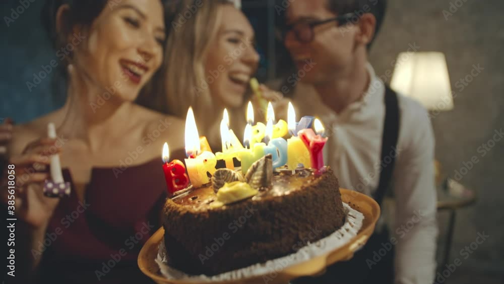 Portrait of asian girl wearing birthday or party cap, blowing whistle and holding birthday cake with burning candles donated by caucasian friends. Happy millennials spending time together at party. Portrait of asian girl wearing birthday or party cap, blowing whistle and holding birthday cake with burning candles donated by caucasian friends. Happy millennials spending time together at party.